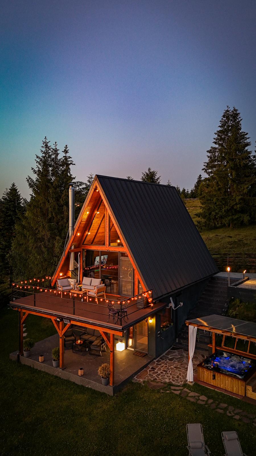 Mountain View Apuseni A-frame cabin at twilight with string lights and hot tub, surrounded by pine trees under a stunning sunset sky